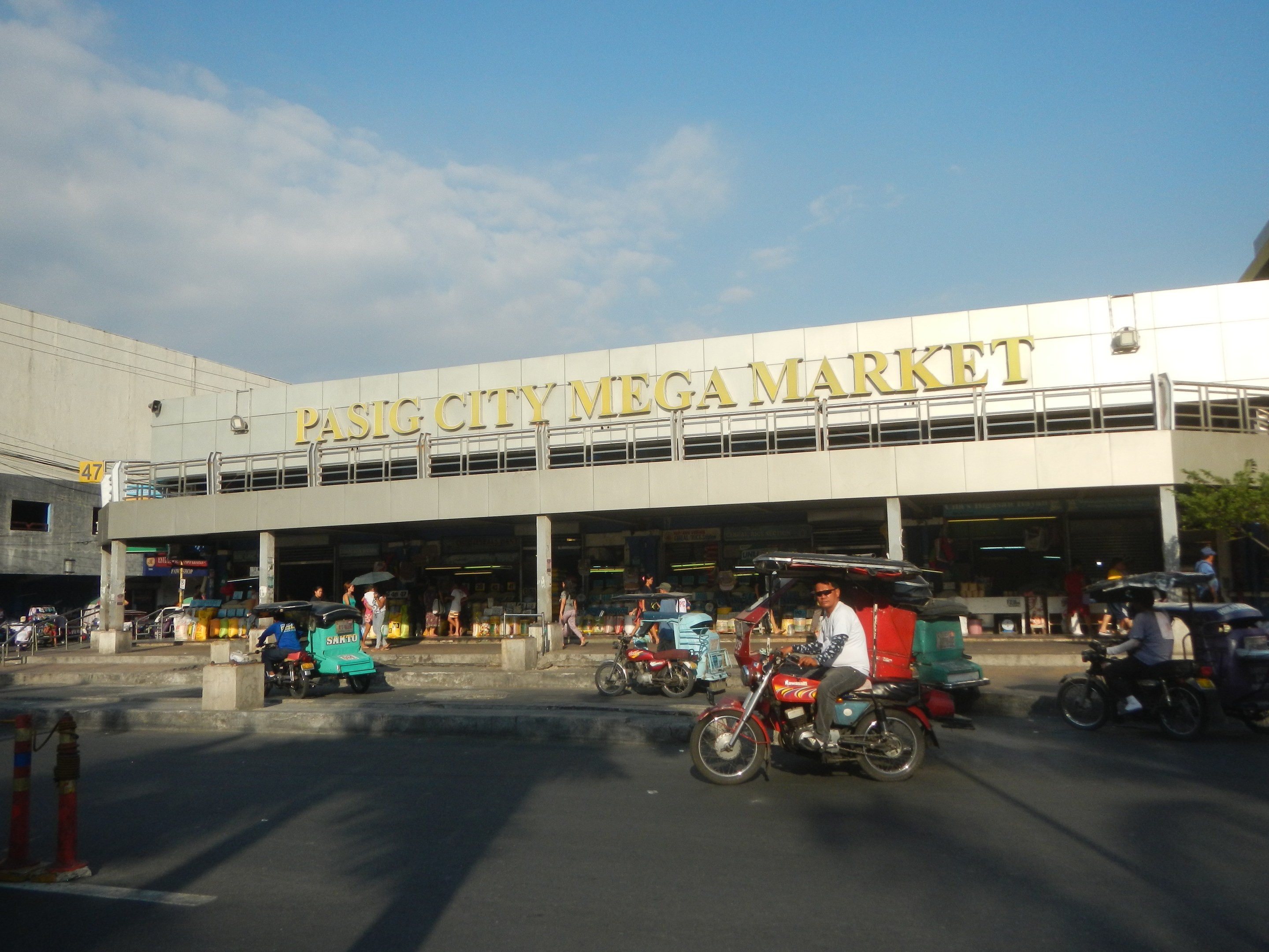 Ein belebter Pasig City Mega Market mit Fahrzeugen auf der Straße, Fußgängern, einem Gebäude mit Text im Hintergrund und einem bewölkten Himmel.