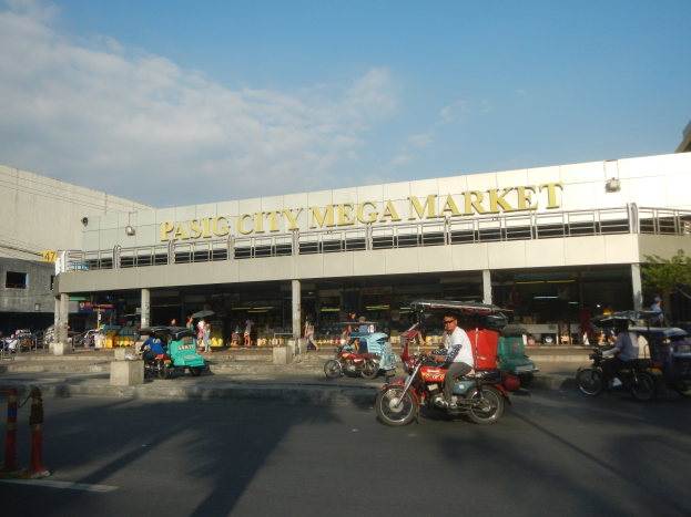 Ein belebter Pasig City Mega Market mit Fahrzeugen auf der Straße, Fußgängern, einem Gebäude mit Text im Hintergrund und einem bewölkten Himmel.