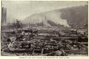 Schwarz-weiß-Foto einer Stadt nach einer Katastrophe am 5. Juni 1892, das Gebäude, Pfähle, Rauch und einen Hügel im Hintergrund zeigt, mit Text am unteren Rand.