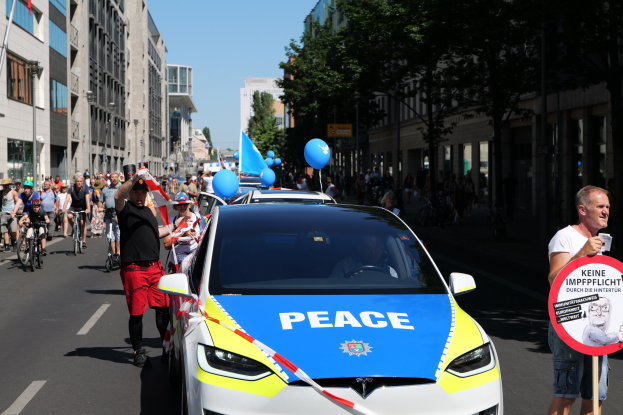 Gruppe von Menschen, die auf Fahrrädern eine Straße entlangfahren, mit einem Polizeiwagen im Vordergrund, einer Person, die ein Schild hält, und Gebäuden, Bäumen und einem klaren Himmel im Hintergrund.