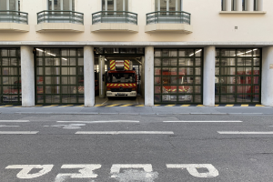 Feuerwache mit vor der Tür geparktem Feuerwehrauto, mit Fenstern, Geländern, Säulen, Glastüren und einer Straße unten.