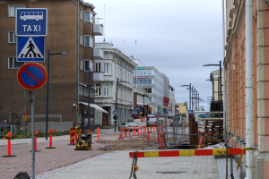 Eine Straßenszene in einer Stadt mit Gebäuden, Fahrzeugen, Verkehrszeichen, Baustelle und einem bewölkten Himmel.