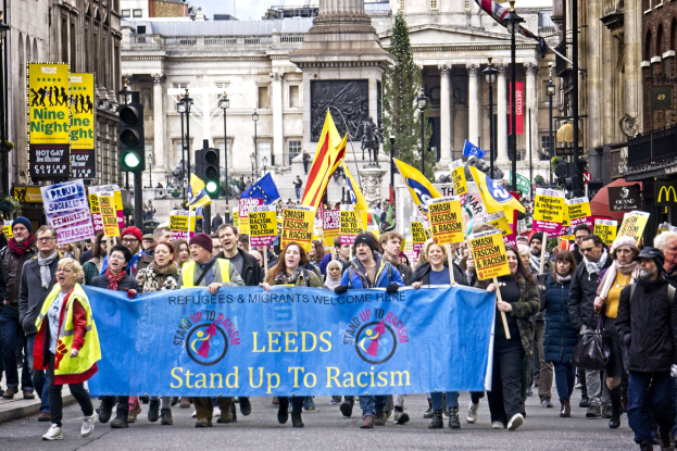 Eine Gruppe von Menschen marschiert mit einer 'Steh Auf gegen Rassismus'-Schlagzeile die Straße entlang, mit Gebäuden, Bäumen, Laternen, Verkehrszeichen und einer Statue im Hintergrund bei bewölktem Himmel.