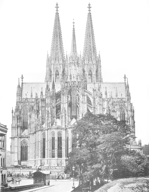 Ein Schwarz-Weiß-Foto des Kölner Doms in Köln, Deutschland, das seine hohen Türme und detaillierte Details zeigt, mit Bäumen, Straßenlaternen, Straßenlaternen und Menschen auf der Straße im Vordergrund und dem Himmel im Hintergrund.