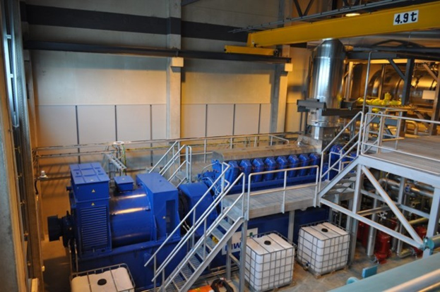 Eine Fabrikinnenszene mit Trägern, einer Treppe, Geländer, Containern, Lichtern, Rohren und Wänden.