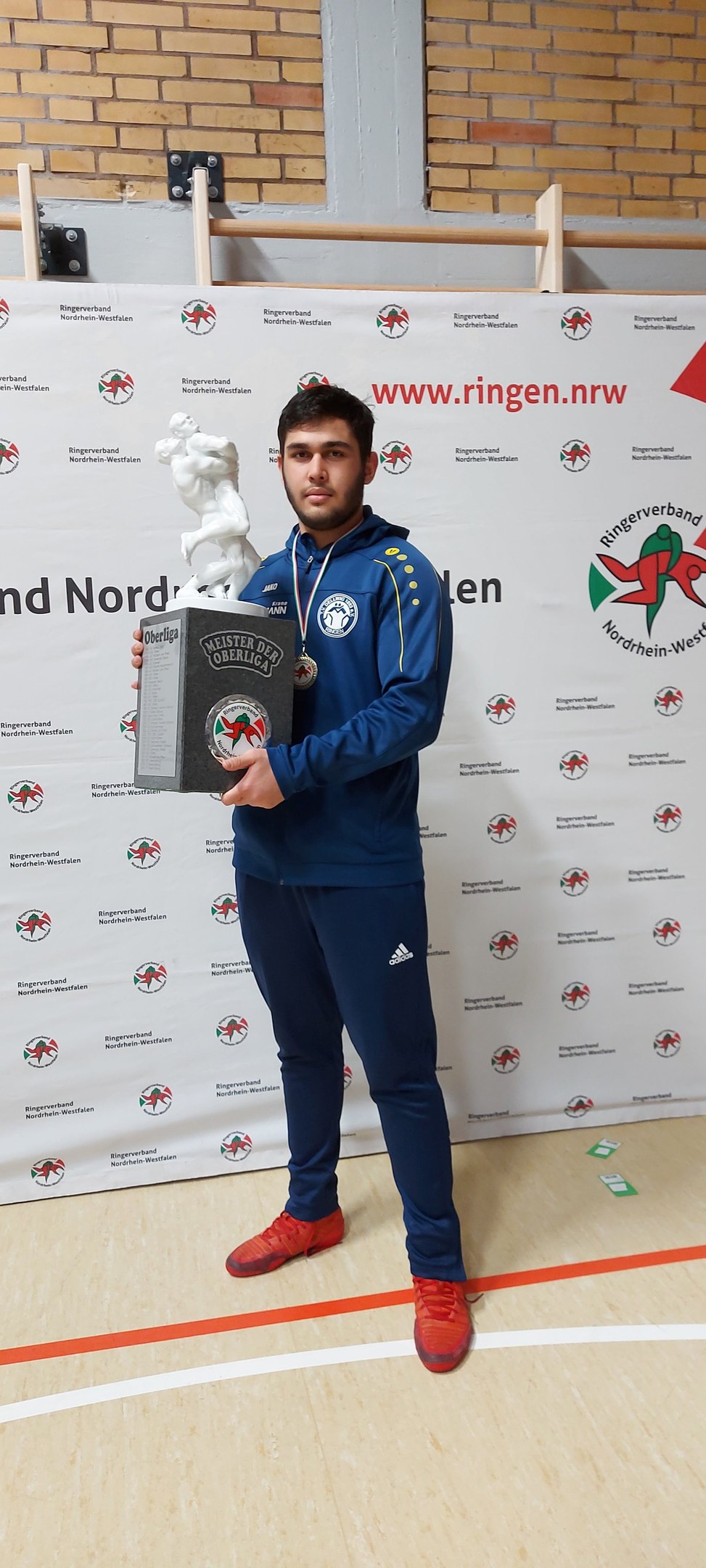Ein Mann in einem Anzug hält einen Pokal, während er eine Medaille trägt, vor einer bannergeschmückten Wand.