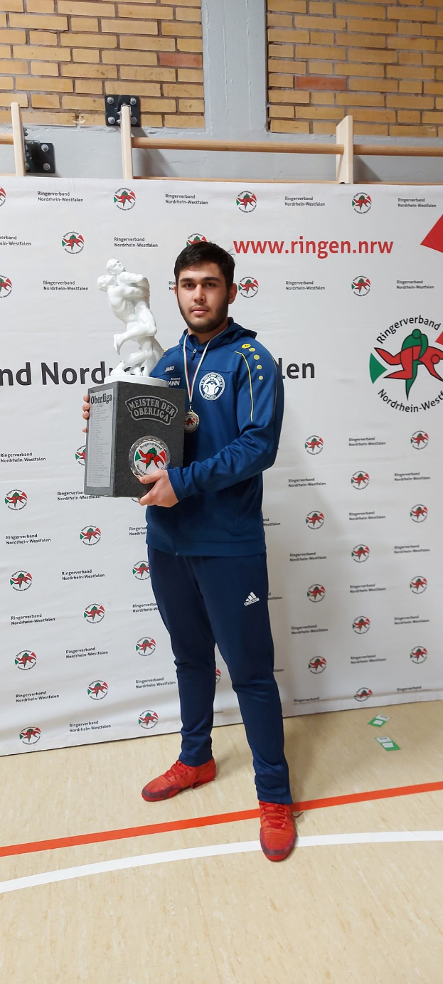 Ein Mann in einem Anzug hält einen Pokal, während er eine Medaille trägt, vor einer bannergeschmückten Wand.