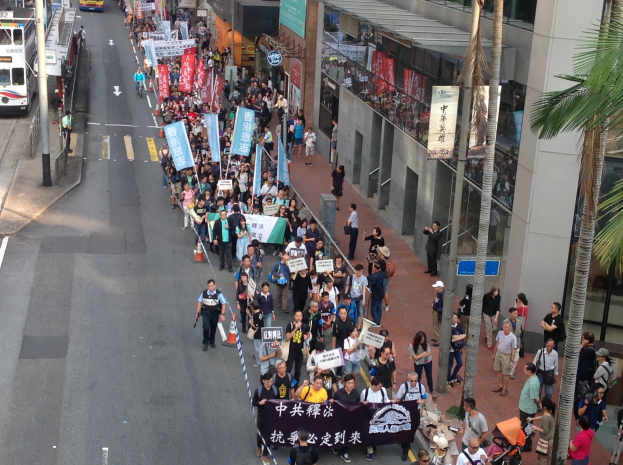 Eine große Gruppe von Menschen marschiert mit Protestschildern die Straße entlang, mit Bäumen und Glasgebäuden auf der rechten Seite und Fahrzeugen auf der linken Seite, was auf eine Protestaktion in Hong Kong hinweist.
