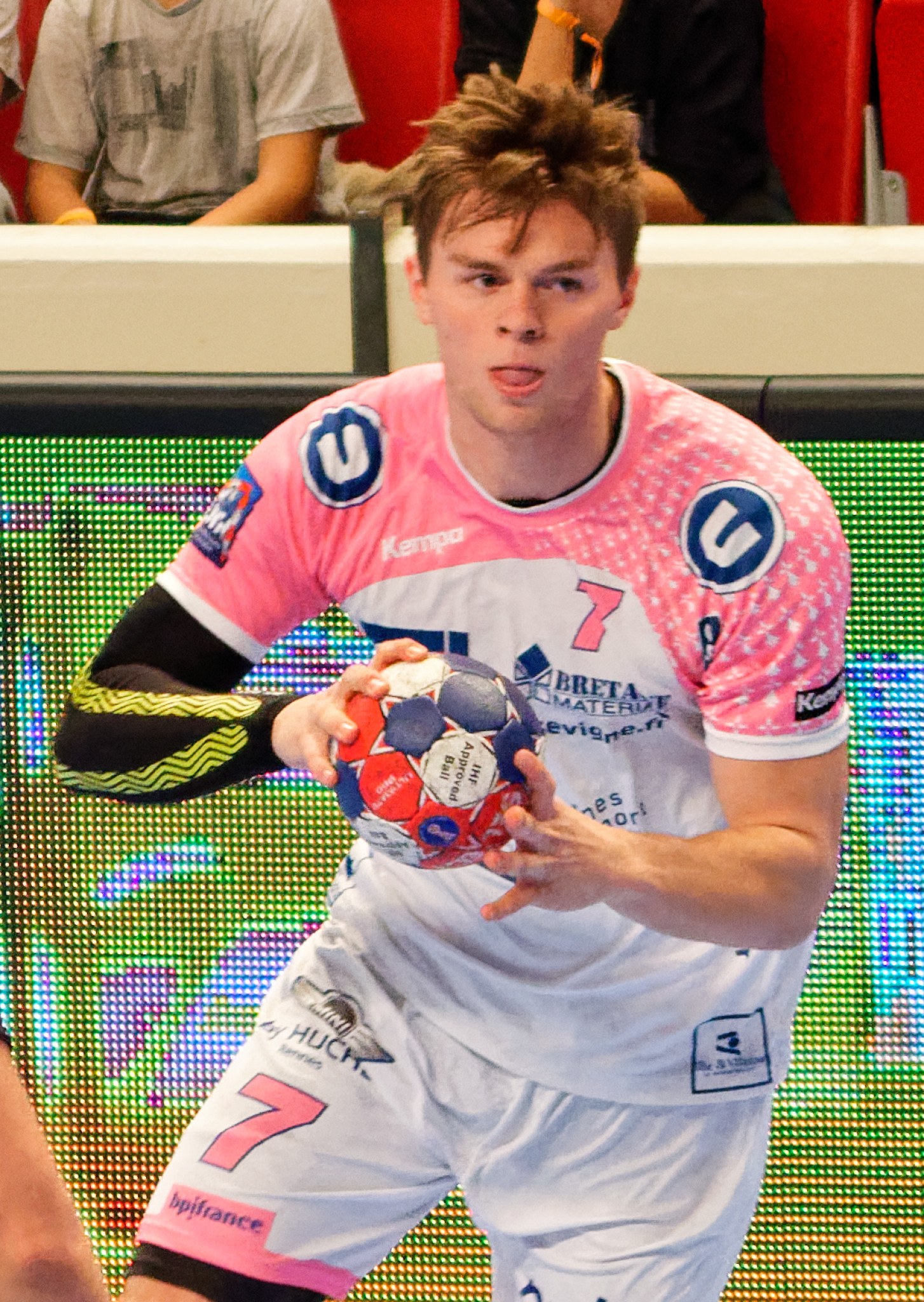 Ein Mann in einem pinken Hemd mit einem Fußball, mit der Hand eines anderen Menschen auf der linken Seite und Menschen im Hintergrund auf Stühlen sitzend.