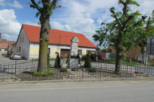 Ein kleines Dorf mit der Kirche des Heiligen Grabes in der Mitte, umgeben von einem Metallzaun, Bäumen, Pflanzen, Gras, einem Schild, Fahrzeugen, Polen, Drähten und einem bewölkten Himmel.