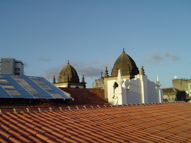 Stadtpanorama mit Gebäuden im Vordergrund, Solarpanelen auf einem Dach und einem blauen Himmel im Hintergrund.