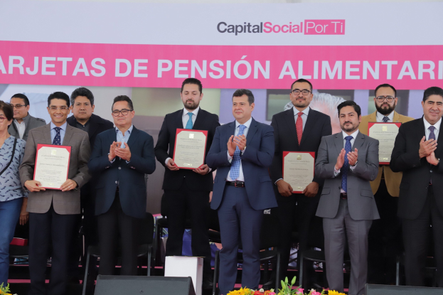 Eine Gruppe von Menschen auf einer Bühne, die klatschen und Zertifikate halten, mit Stühlen im Halbkreis drumherum, einem Banner mit der Aufschrift "Capital Social for Ti" im Hintergrund und Blumensträußen und Lautsprechern unten.