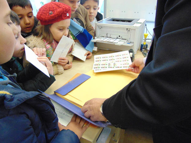 Mehrere Kinder halten Papiere in den Händen, ein Mädchen trägt eine Mütze, während ein Mann in der Nähe Papiere in einer und Umschläge in der anderen Hand hält, mit einem Drucker auf dem Tisch.