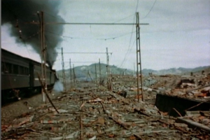 Ein Zug fährt durch eine geschädigte Gegend mit Rauch, der aus ihm aufsteigt, umgeben von Strommästen und -drähten, mit Höhen und einem klaren Himmel im Hintergrund und verstreuten Trömmern am Boden.