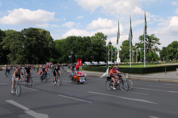 Eine Gruppe von Menschen, die auf Fahrrädern eine von Bäumen gesäumte Straße mit Laternenmasten und Fahnenmasten entlangfahren, während sie an der Pariser Fahrradfahrt 2016 teilnehmen.